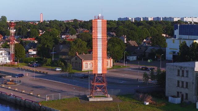 Aerial view over Ventspils shows an orange and white daymark on an iron base by the quay, the Vizium center, homes, cars on the road, and pedestrians by the water.