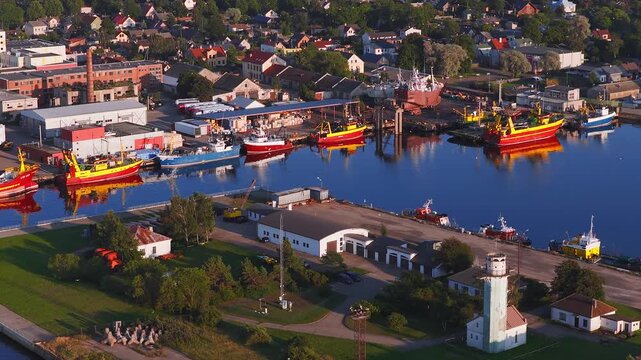 Aerial view of Ventspils, Latvia port shows white lighthouse, cranes, brick chimney, warehouses, homes, and moored trawlers with calm water reflections at low light.