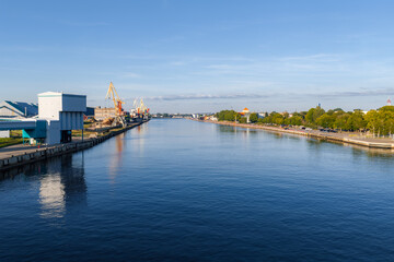 Naklejka premium Wide channel toward the Port of Ventspils in Latvia, cargo cranes and terminals on the left, a promenade on the right, calm water in morning light, modern buildings and silos.