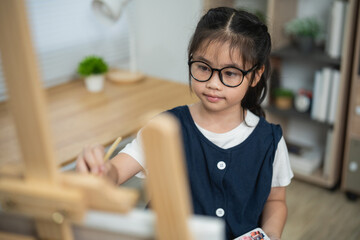 Focused Asian Little Girl with Glasses Painting on Canvas with Wooden Easel and Holding Paint Palette at Home Studio © WMSTUDIO
