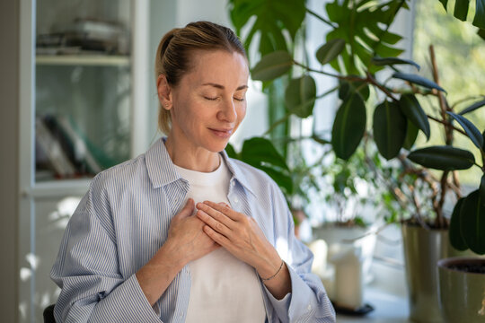 Relaxed calm woman meditates with hands on chest, practicing breathing exercises at home. Mindfulness practice, focus on breath, self care routine, stress relief, reflection and holistic wellness