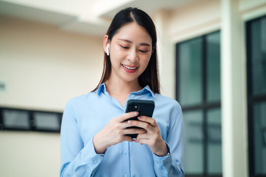 Happy Asian businesswoman using smartphone with wireless earbuds standing outside office building