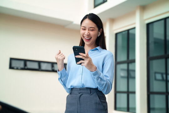 Excited Asian businesswoman celebrating success with fist pump while looking at smartphone