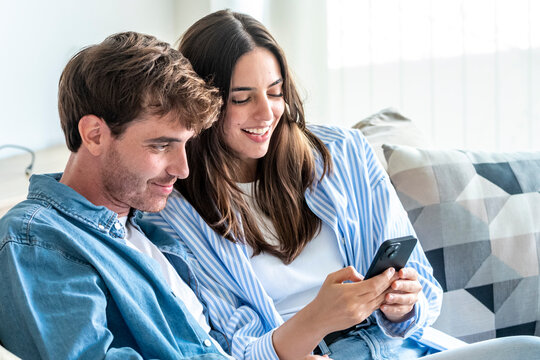 Young couple sitting comfortably on a sofa at home, smiling while sharing and looking at content on a mobile phone, representing connection, leisure, and modern lifestyle