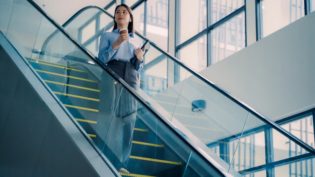 Asian businesswoman with tablet and coffee cup riding escalator inside modern building