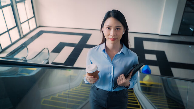 Confident Asian businesswoman holding tablet and coffee cup looking at camera on escalator