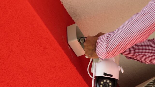 Professional electrician mounts and secures a CCTV security camera onto a wall-mounted junction box using a cordless drill.