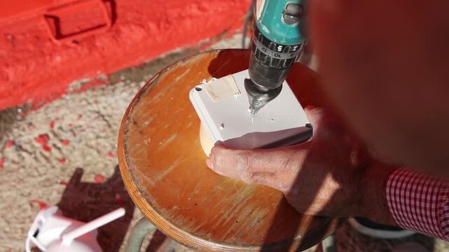 Close-up of an electrician drilling a cable entry hole in a plastic junction box while preparing an outdoor CCTV security camera installation.