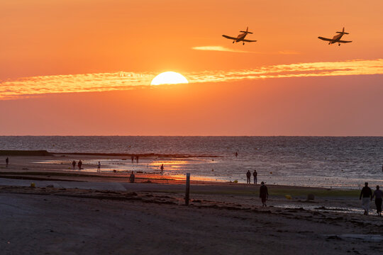 View the beach, sea with people walking, two single-engine propeller-driven light aircraft flying in the sky and the sun low on the horizon at golden hour with clouds