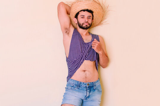 Man with a fisherman-style look and hat, unbuttoning his shirt. Man in summer attire, fisherman with a straw hat. Young man wearing a blue striped tank top and denim shorts light clothing, summer