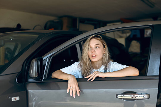 Young woman leans out of car window in a garage during daytime while looking lost in thought