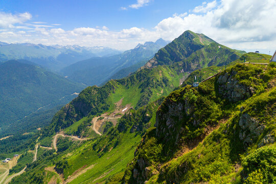 Panoramic view of green mountain peaks under a blue sky in Sochi, Russia. Winding road on a steep grassy slope in a high altitude landscape. Summer travel, nature and adventure