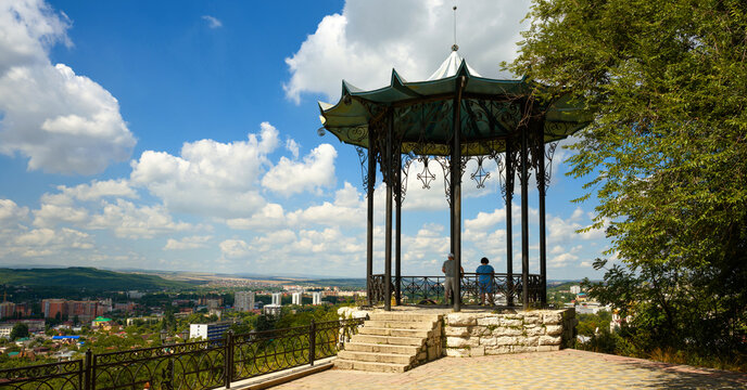 Ornate wrought-iron gazebo on a stone platform overlooking a Pyatigorsk city, Russia. Tourists standing in a pavilion enjoying the landscape view. Summer sightseeing destination under a blue sky