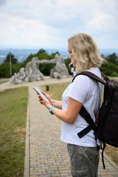 Vertical photo of a blonde woman tourist using a smartphone. Female traveler with a backpack standing on a paved path in Pyatigorsk, Russia. Outdoor navigation and travel concept