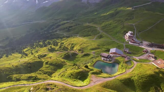 Aerial drone view of Kleine Scheidegg in Swiss Alps with green meadows and snowy Eiger Monch peaks in early morning light. Small station with emerald lake and winding road in 4K video
