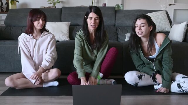 Three young women sitting in a lotus position in their living room, following an online yoga class on a laptop, beginning their meditation and mindfulness practice together at home