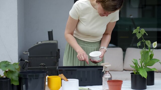 Woman sows seeds into black planter box while holding bowl at patio gardening table. Concept of container gardening and plant care