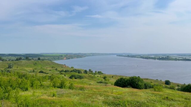 Wide river flows gently under clear sky. Grassy hillside sways with wildflowers in breeze. Distant trees frame serene water surface. Nature's quiet beauty captured in calm, open view.summer. The Kama 