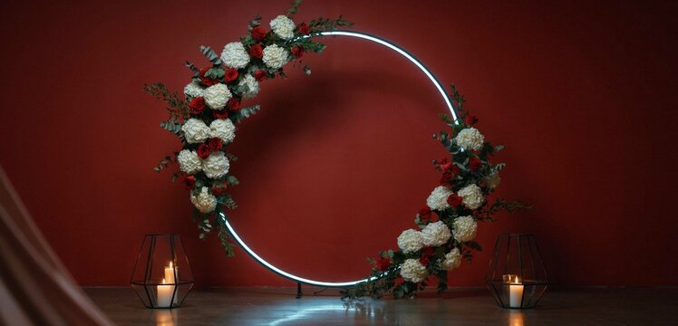Modern round glowing neon wedding arch decorated with white hydrangeas and red roses against dark red background