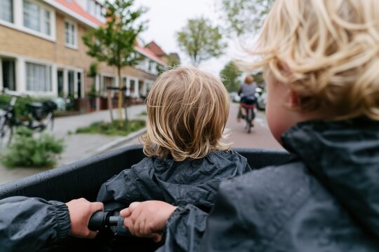 two young children with blonde hair riding in a cargo bike on a rainy street
