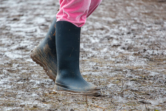 The girl is wearing pink pants and dark rubber boots. The boots are dirty, and she walks on wet, frozen ground.