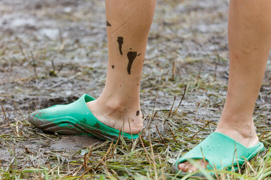 Person wears green slippers in muddy field. Mud splatters their ankles and lower legs.