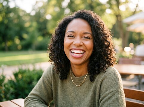 Happy young african american woman with curly hair smiling brightly while sitting at an outdoor cafe table in a green park