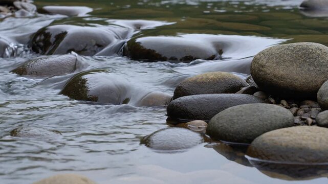 Clear mountain stream water flowing over smooth river rocks creating a tranquil and relaxing natural scenery. The serene current creates a calming meditation background