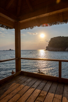 Wooden seaside pavilion with shingled roof and railing overlooking rocky cove, calm blue water, distant hills, and warm golden hour light across coastal floorboards
