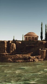 A structure stands on a rocky edge overlooking water. Tall trees rise beside the building. The scene shows a clear blue sky and calm water. The area appears untouched and natural.