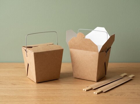 Two brown cardboard takeout boxes for asian food with wooden chopsticks on a wooden table