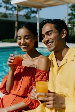 Smiling young couple holding orange and yellow drinks by a pool for summer lifestyle banner and vacation background