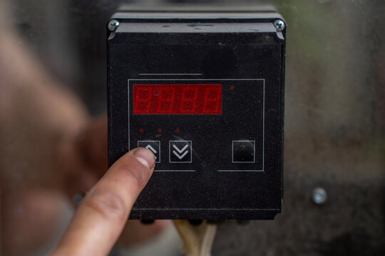 Finger pressing arrow up button on digital temperature controller panel with red led display screen in industrial setup.
