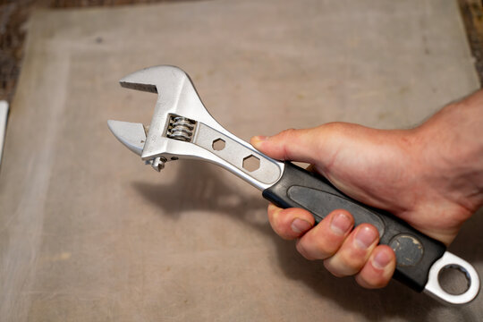 Male hand holding an adjustable spanner wrench over a workbench, showing manual repair work, plumbing maintenance, mechanical service, and hand tool operation.