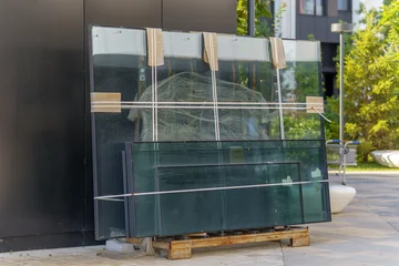 Glass panes packed on a metal pallet stored outdoors near a building facade. Large window units and double glazed panels secured with straps before construction installation. © sandipruel