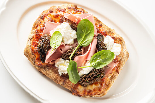 pinsa romana pizza with mortadella, stracciatella cheese, morel mushrooms, and fresh spinach on a white plate.
