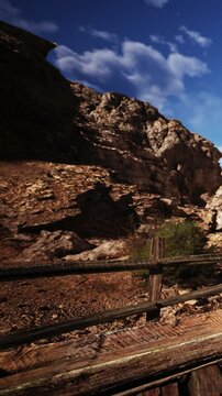 A wooden fence stands in front of rocky terrain and steep cliffs. Bright sunlight illuminates the mountains under a blue sky with a few clouds. This scene shows natures raw beauty.