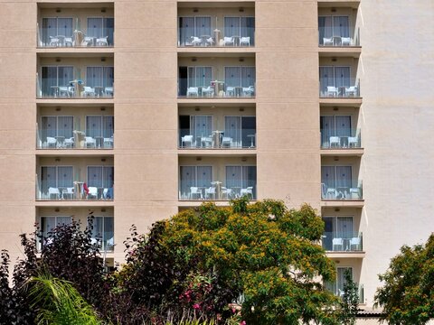 Front view of a multi-story hotel facade displaying symmetrical rows of room balconies with glass railings, framed by lush green and purple trees in full sunlight.