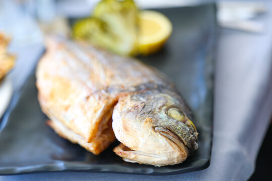 Whole grilled fish served on a black plate with lemon and vegetables, showcasing fresh Mediterranean cuisine and traditional seafood dining.