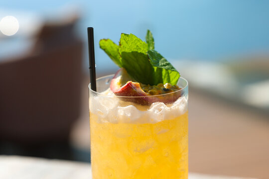 A tall glass of tropical cocktail garnished with mint and citrus sits beside a pool overlooking the Aegean Sea in Greece, glowing in golden Mediterranean sunlight.