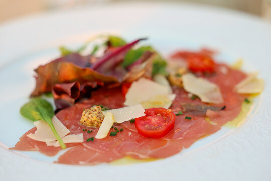 Thin slices of beef carpaccio topped with cherry tomatoes, parmesan shavings, mustard, and mixed greens, plated elegantly in a fine Greek restaurant.