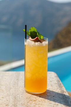 A tall glass of tropical cocktail garnished with mint and citrus sits beside a pool overlooking the Aegean Sea in Greece, glowing in golden Mediterranean sunlight.