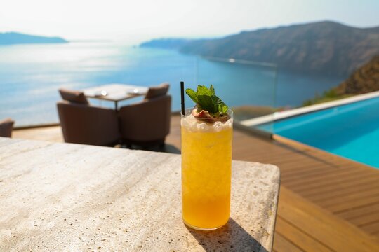 A tall glass of tropical cocktail garnished with passionfruit, mint and citrus sits beside a pool overlooking the Aegean Sea in Greece, glowing in golden Mediterranean sunlight.
