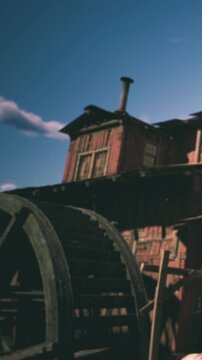 A large water wheel stands next to a rustic wooden building. The structure shows signs of age. The sky is clear with a few clouds. Sunlight highlights the features of the scene.