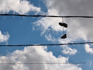 Obraz premium Old sneaker pair hanging on electric wire against blue sky with clouds. Shoefiti concept on utility cable.