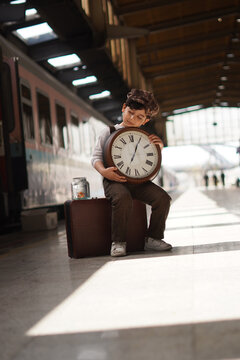 Vintage dressed boy holding large clock on suitcase at train station