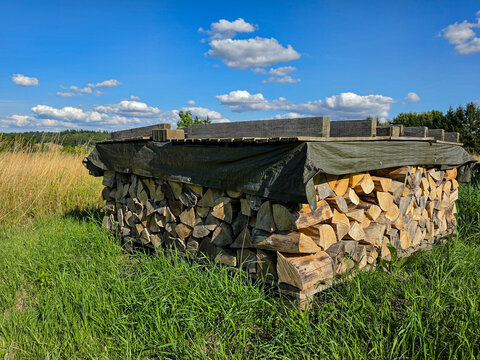 stacked firewood covered with protective tarp in a rural meadow under a blue summer sky