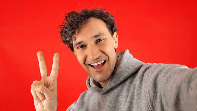 Happy young man with curly hair in grey hoodie taking selfie, making peace sign gesture against solid bright red background expressing positivity success lifestyle.
