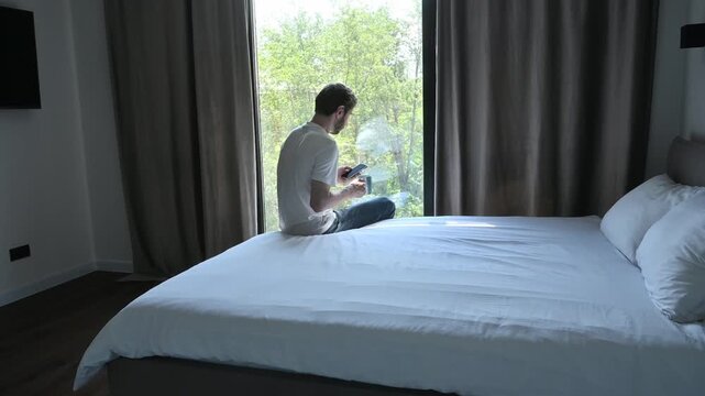 Man sitting on the bed, using his smartphone and drinking coffee in the morning