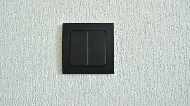 Black dual light switch on textured white wall, male hand pressing it, turning the lights on